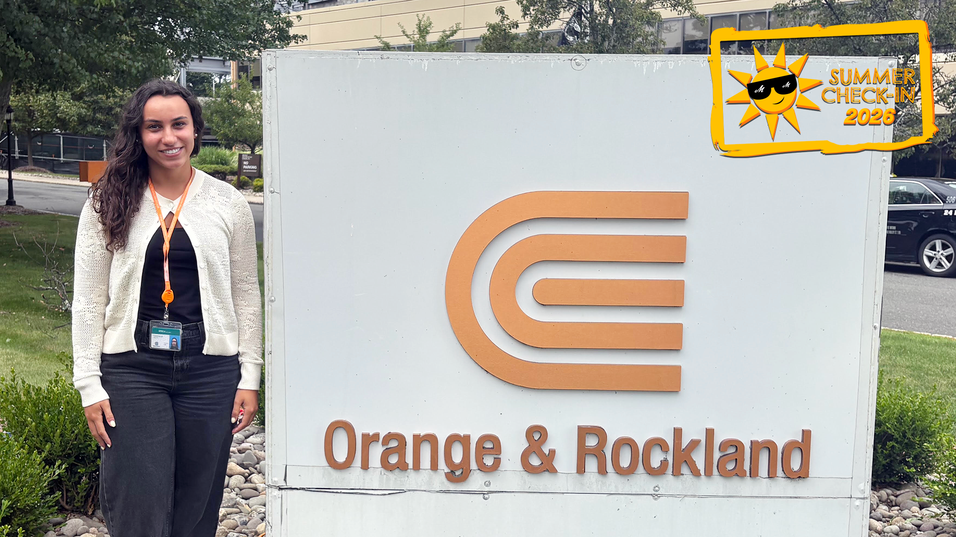 a young woman wearing a black outfit and white sweater standing outdoors next to a white slab that says Orange & Rockland in orange