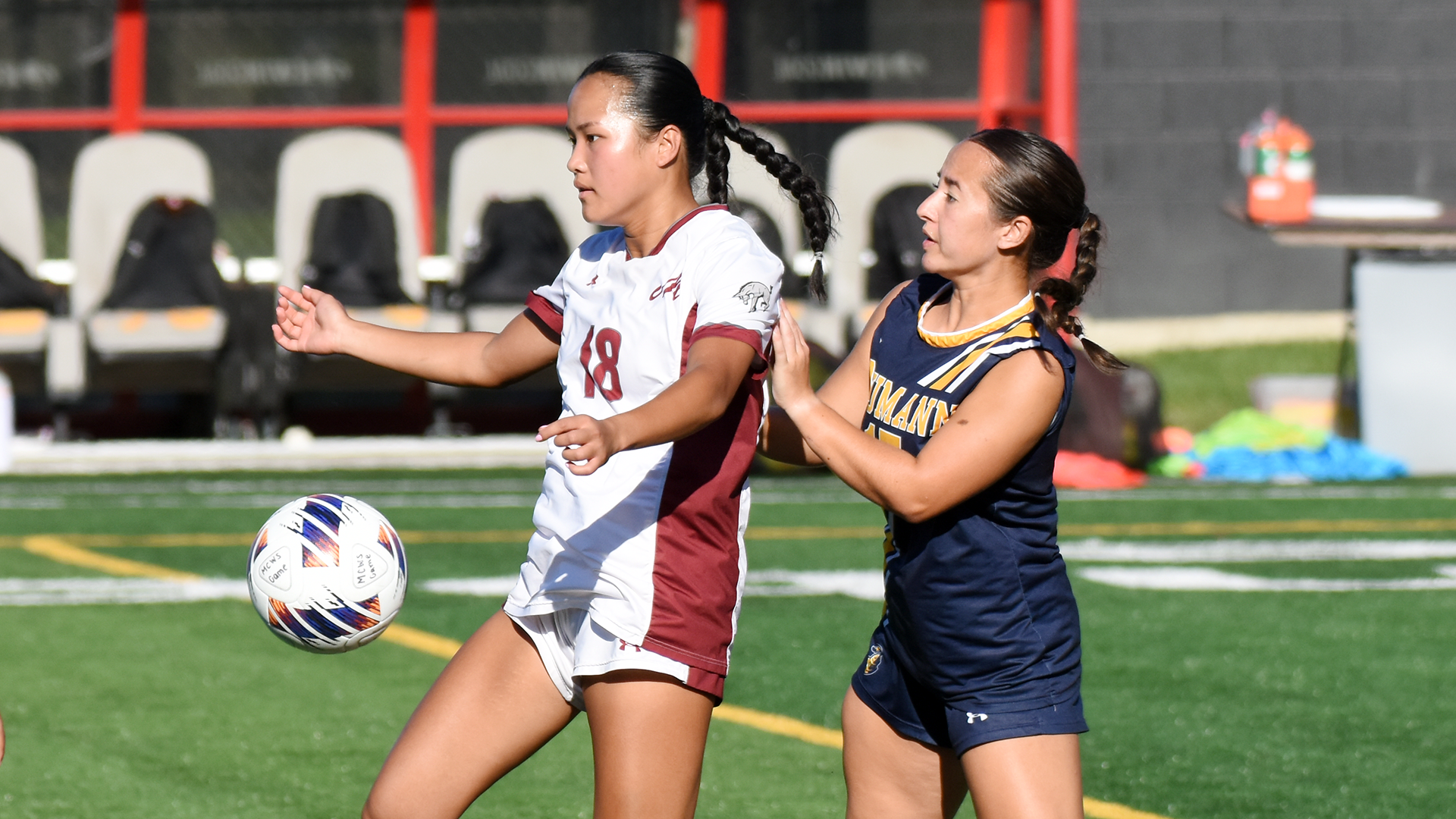 a women's soccer player in a white uniform controls the ball on a sunny day with a player in a blue uniform behind her