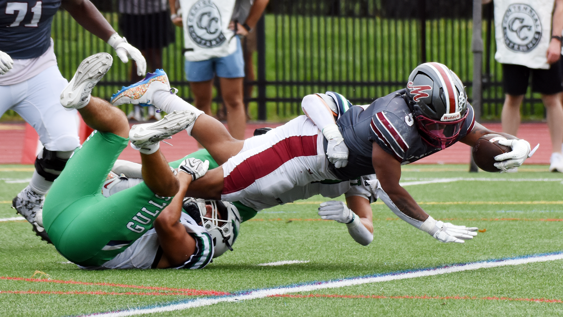 a football player in a grey shirt, grey helmet and white pants stretching across the goal line while a player in a white shirt and green pants holds his legs