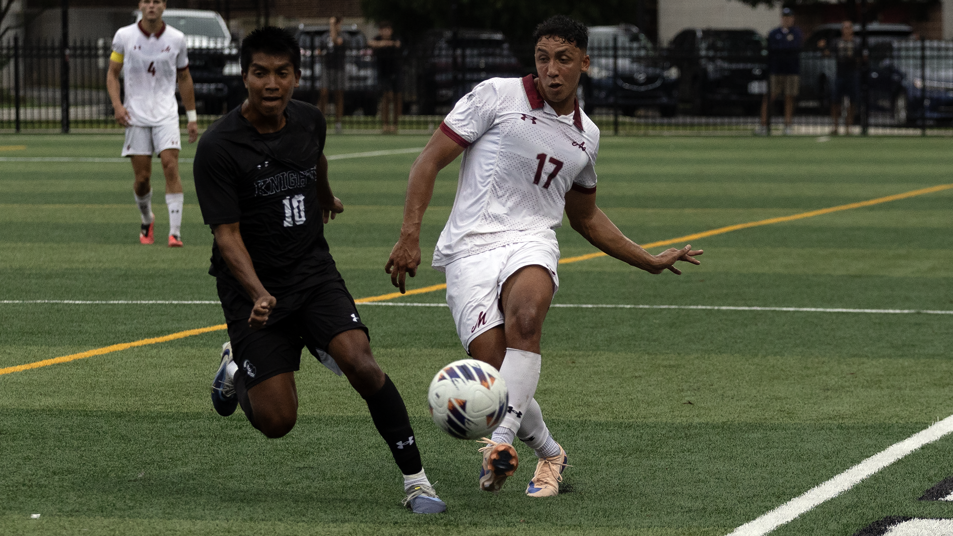 a men's soccer player in a white uniform kicking the ball with his left foot as a player in a black uniform closes in