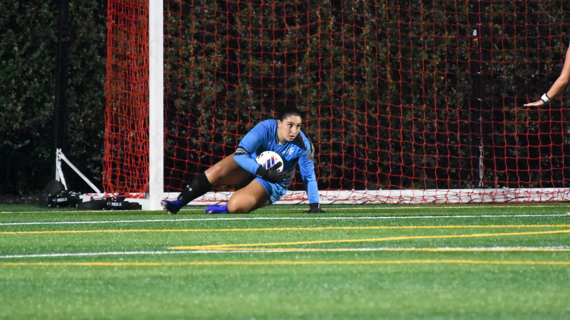 A goalie in a blue uniform gets up off the ground in front of the goal while holding the ball after making a save