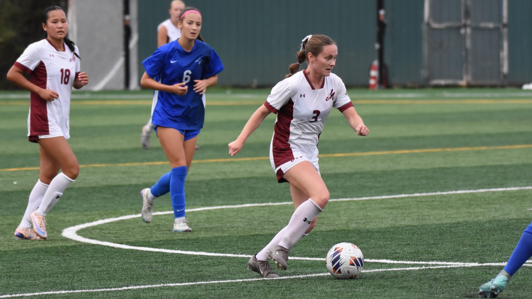 A soccer player in a white jersey dribbles the ball upfield during a women's soccer game