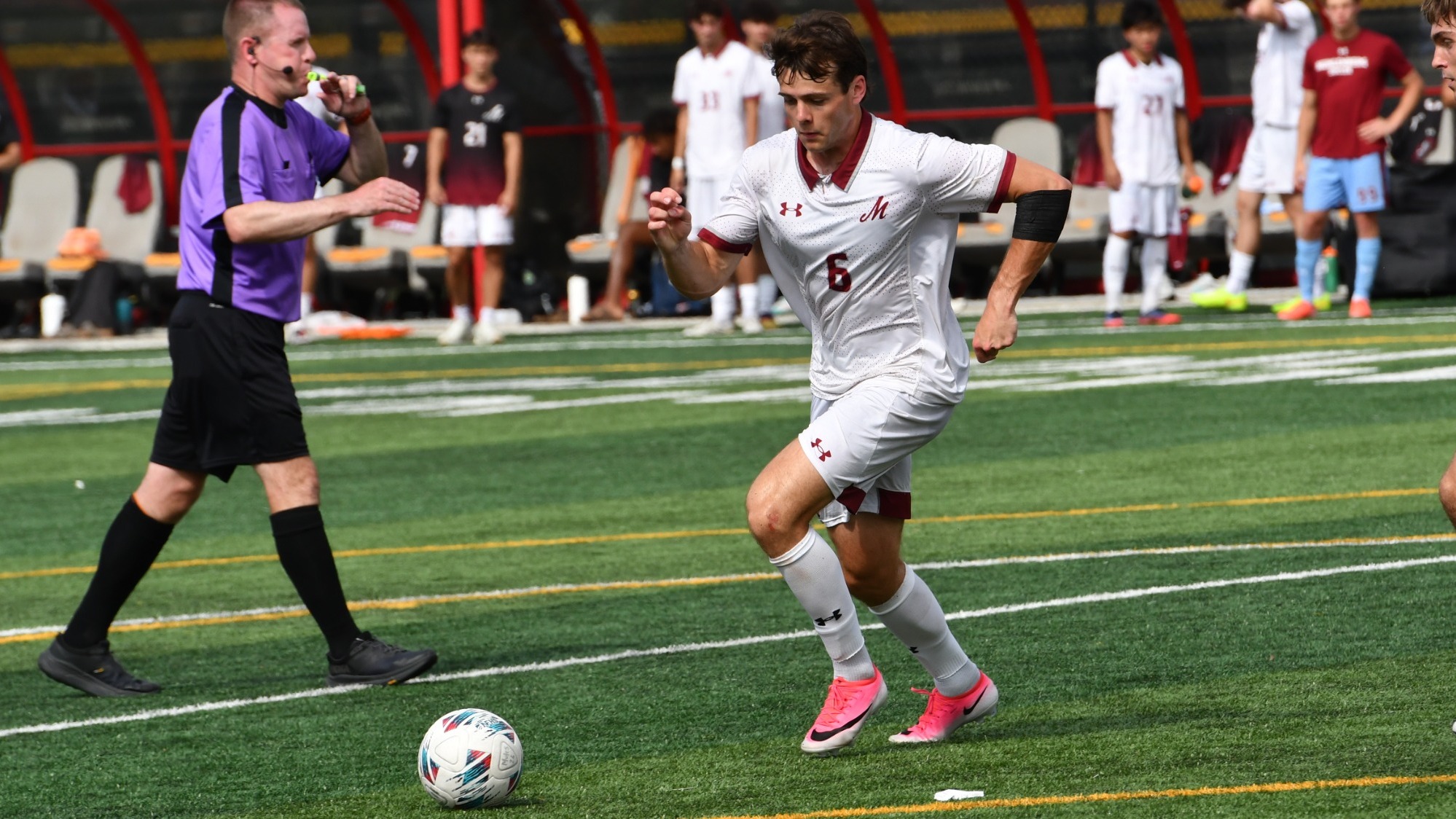 A Muhlenberg soccer player in a white uniform runs with the ball during a game as a referee follows nearby.