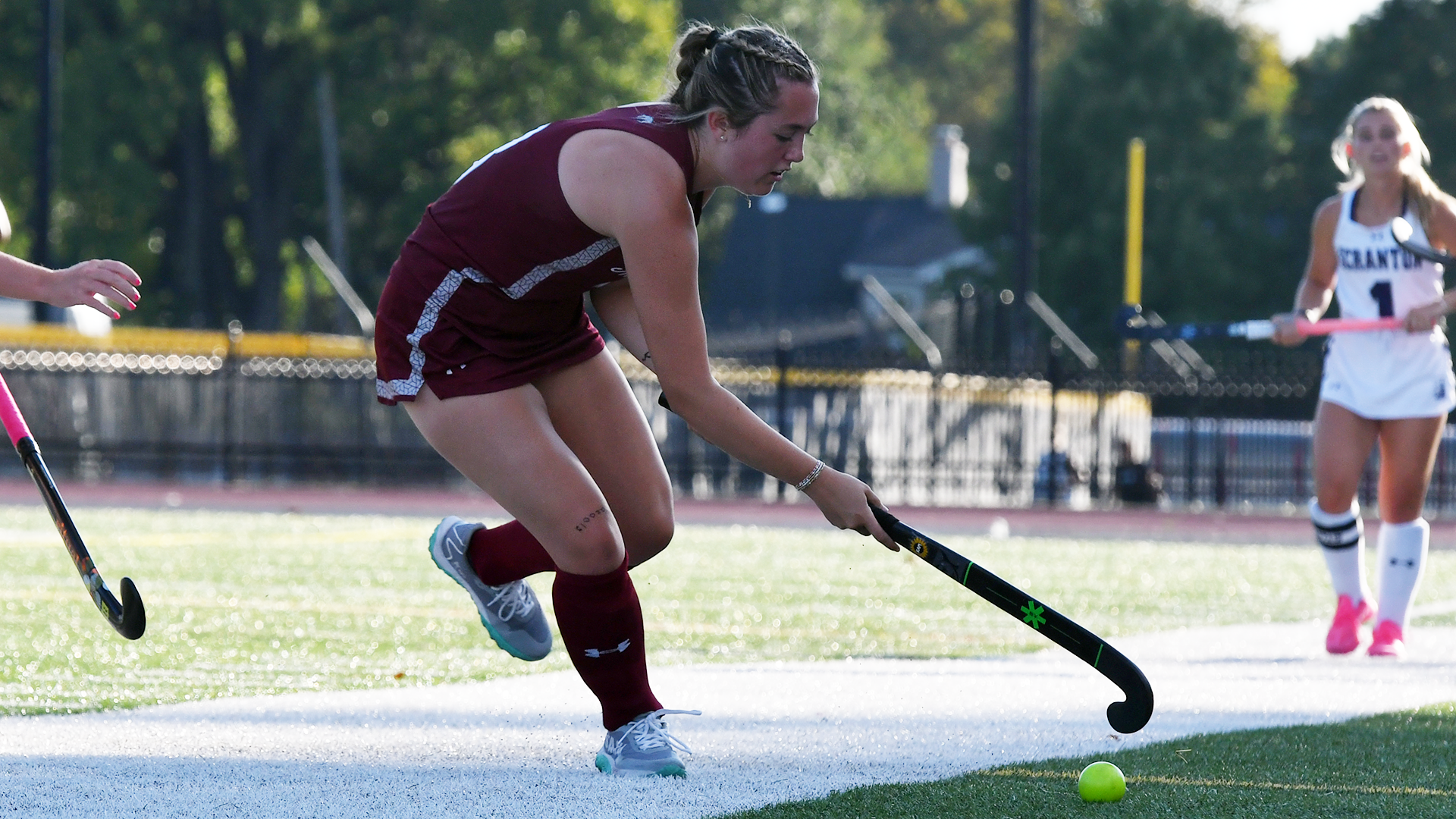 a field hockey player in a red uniform facing right with her stick extended to the ground to hit the ball