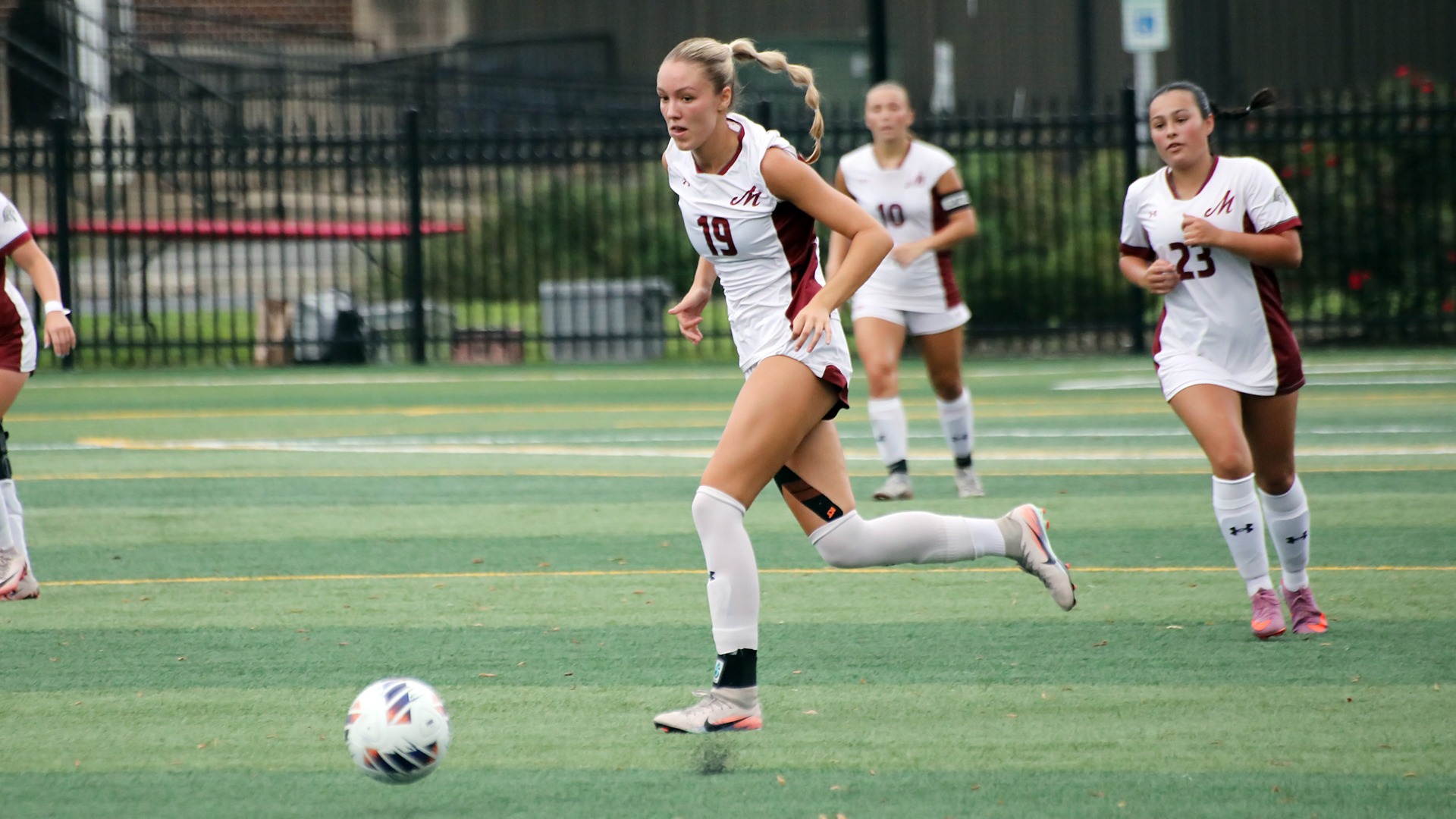 a soccer player in a white unform dribbling on a green turf field with teammates behind her