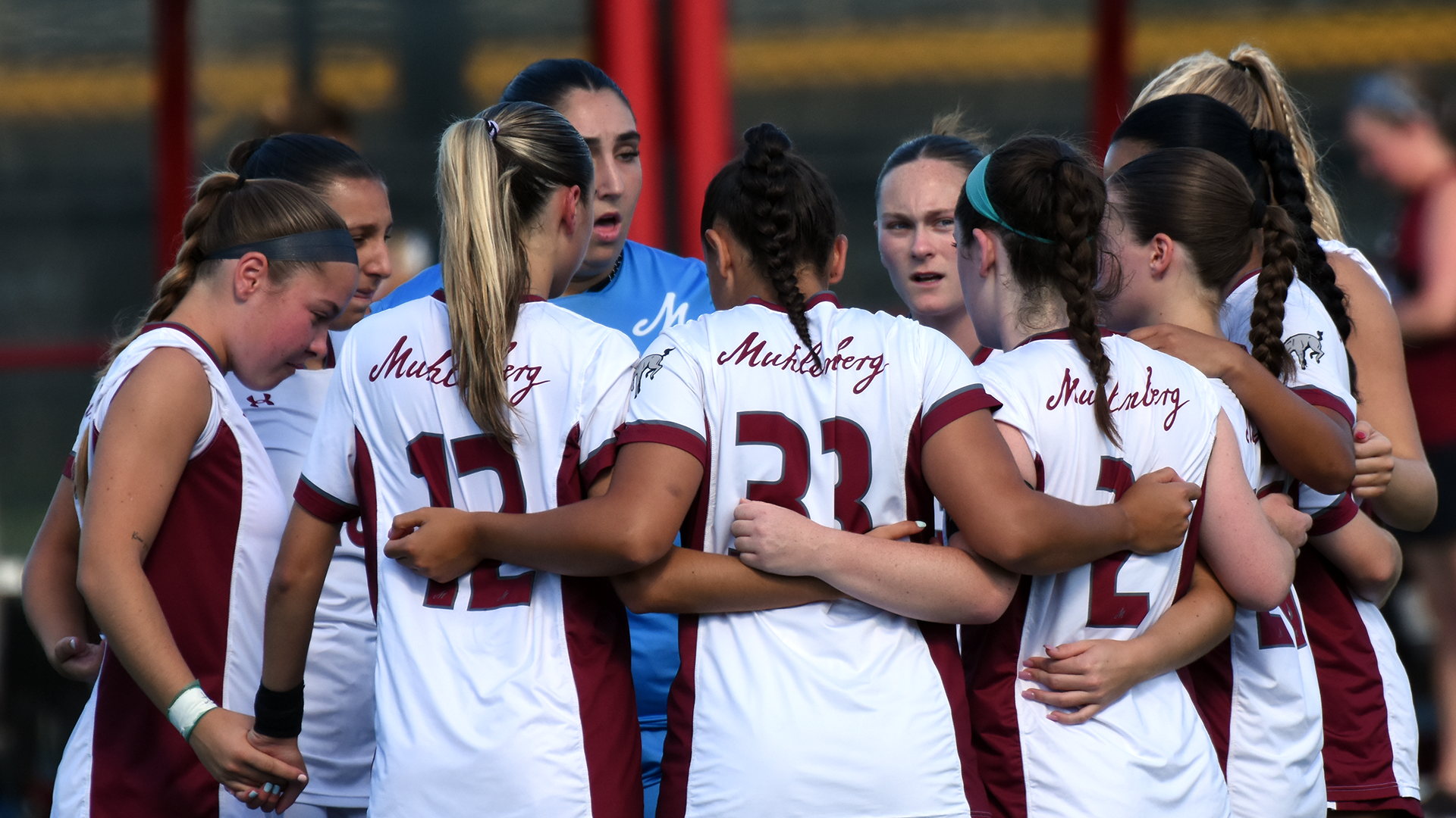 women's soccer players in white uniforms stand in a circle on a sunny day