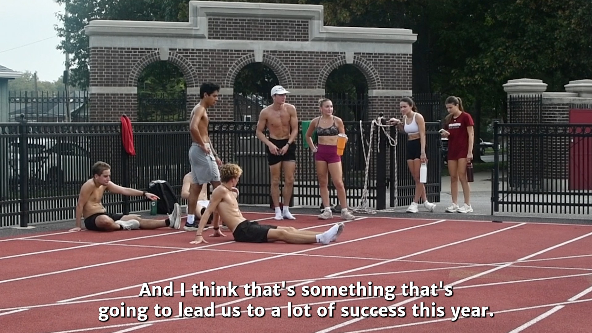 cross country runners stretching and standing on a red outdoor track with iron and brick gates behind them