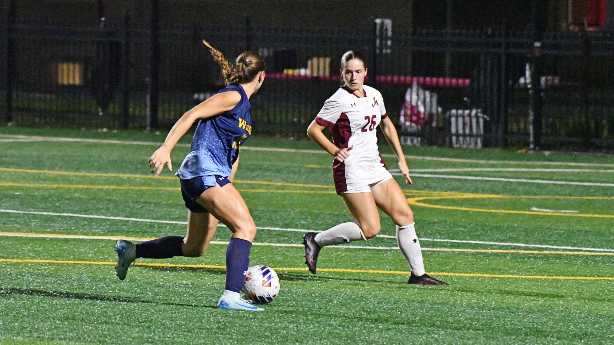 A soccer player in a white and maroon uniform runs alongside an opposing player in blue while competing for the ball.