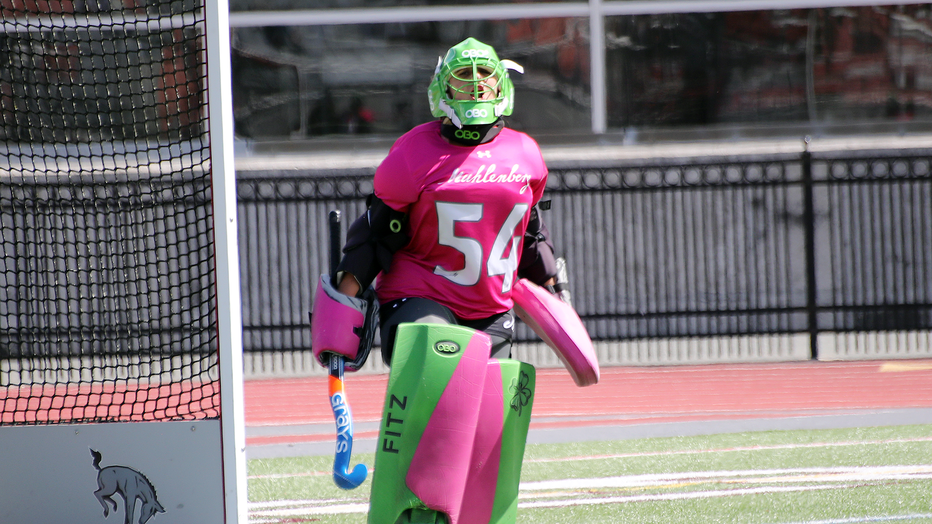 a field hockey goalie in a pink shirt and green pads yelling at her teammates on a sunny day