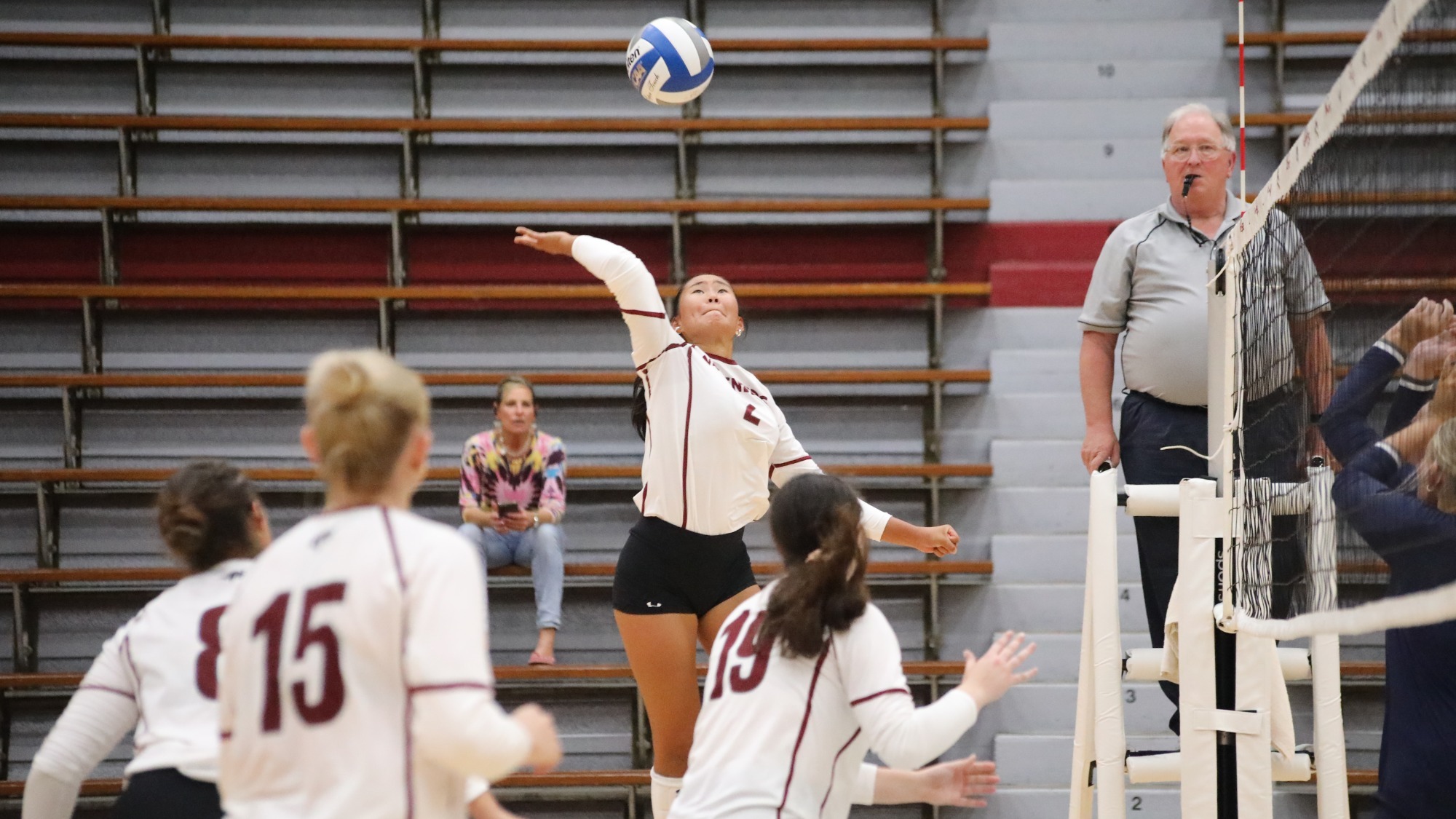 A volleyball player in a white uniform jumps to hit the ball over the net as teammates and an official watch the play.