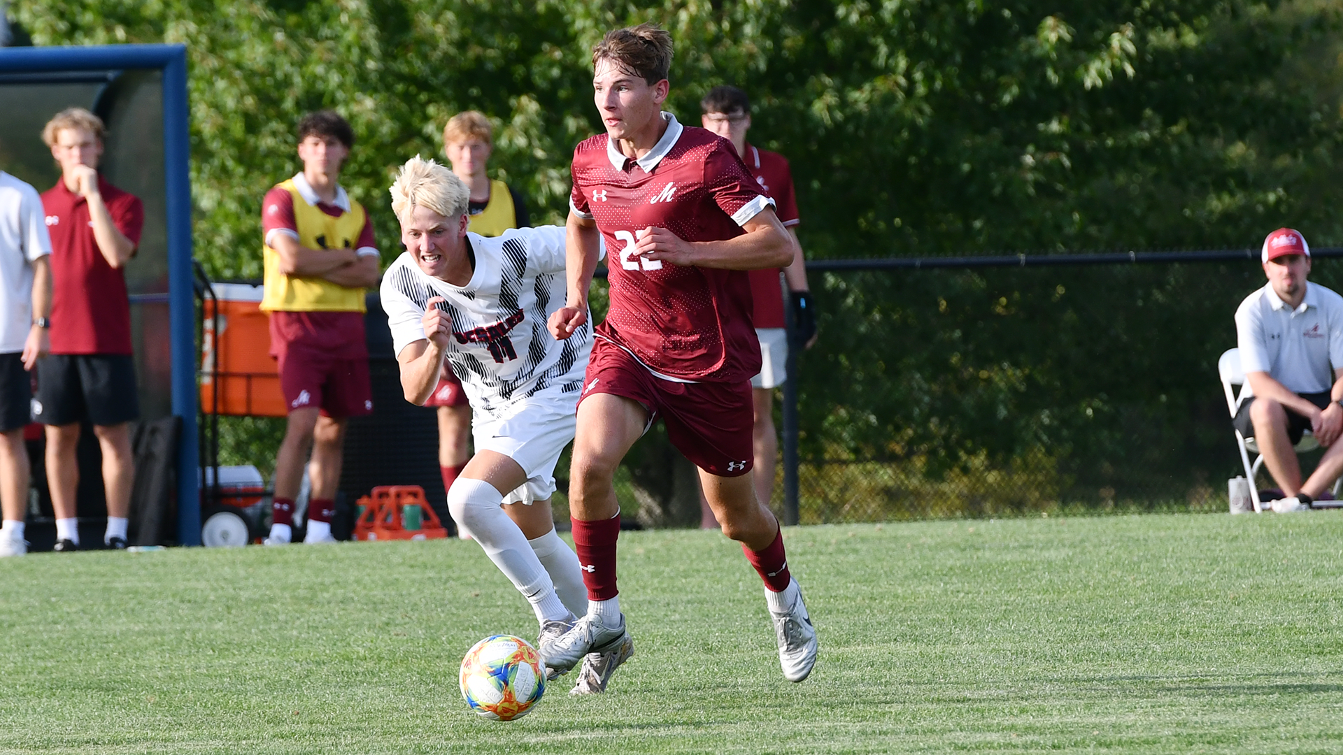 a soccer player in a red uniform dribbling on a grass field while being guarded by a player in a white uniform