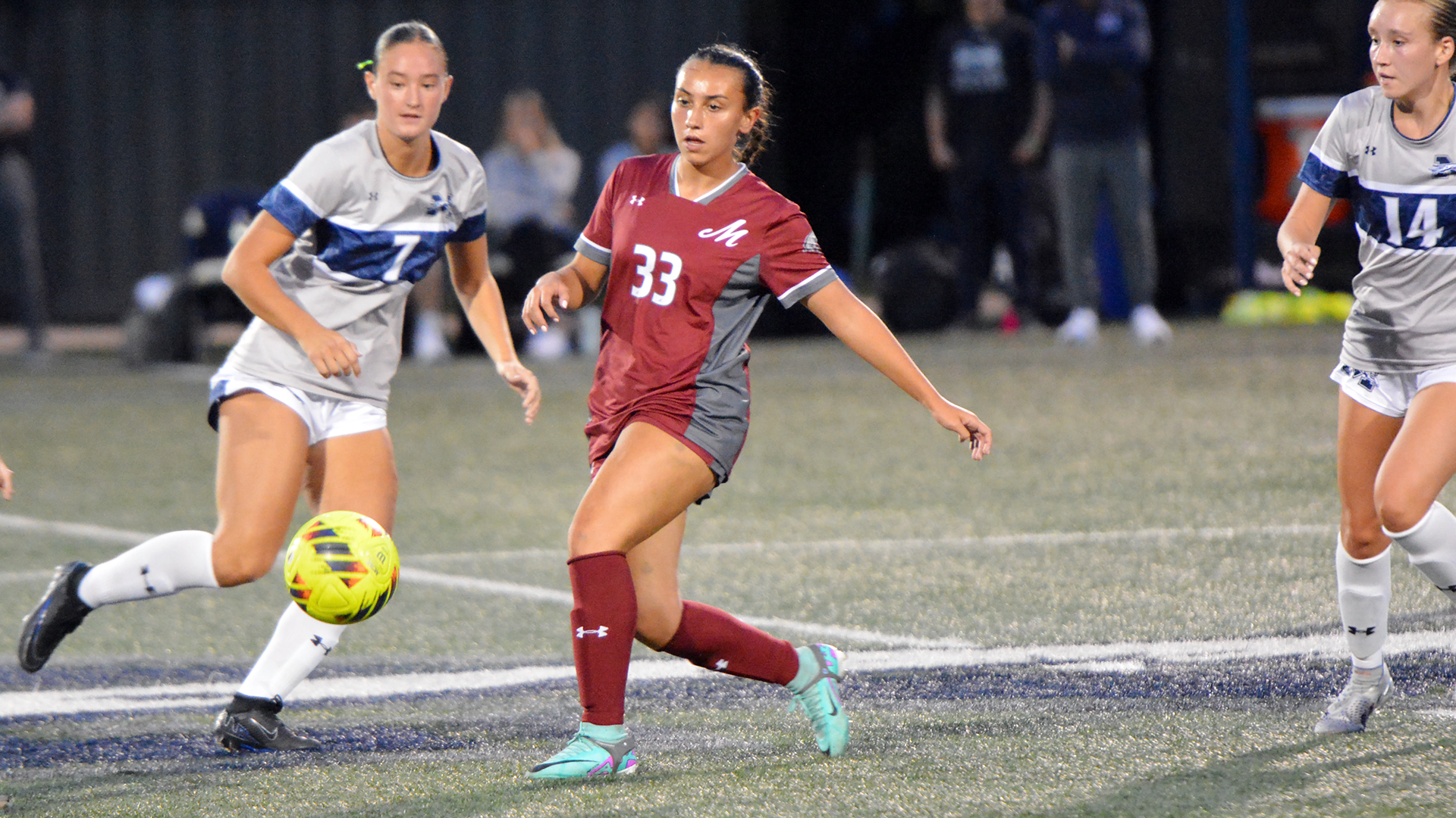 a women's soccer player in a red uniform passing in between two players in blue and grey uniforms at night
