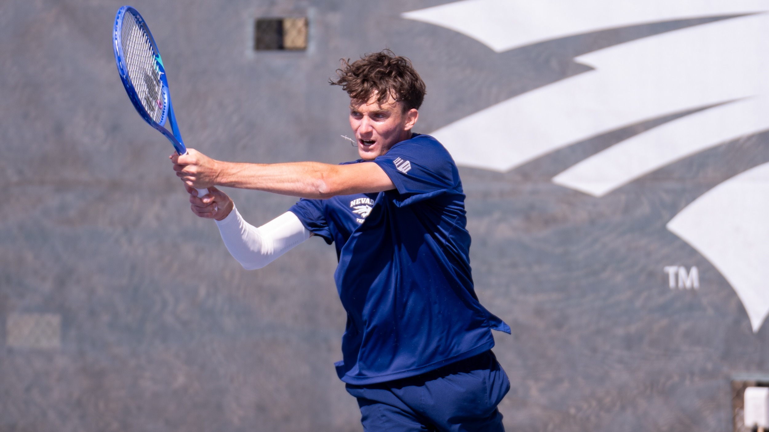 A Nevada menb's tennis player in a navy blue shirt in his backhand