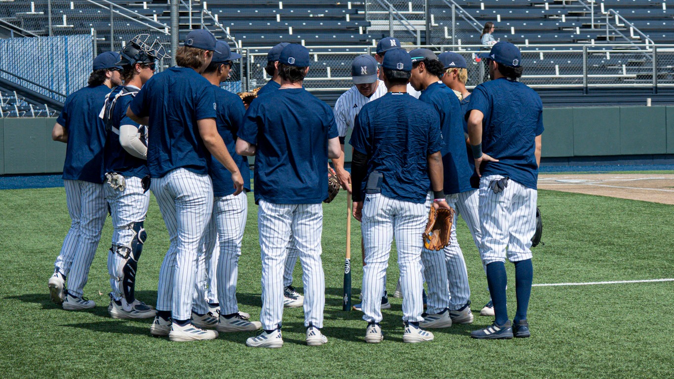 A group of Nevada baseball players in navy warmup tops in a huddle by the dugout