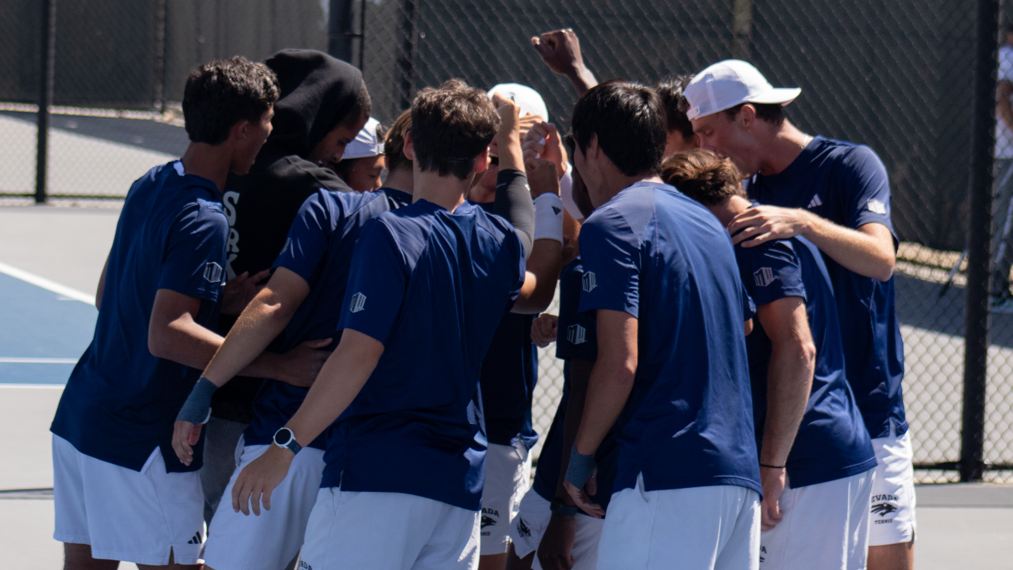 Nevada Men's Tennis team in navy blue shirts and white shorts in a team huddle
