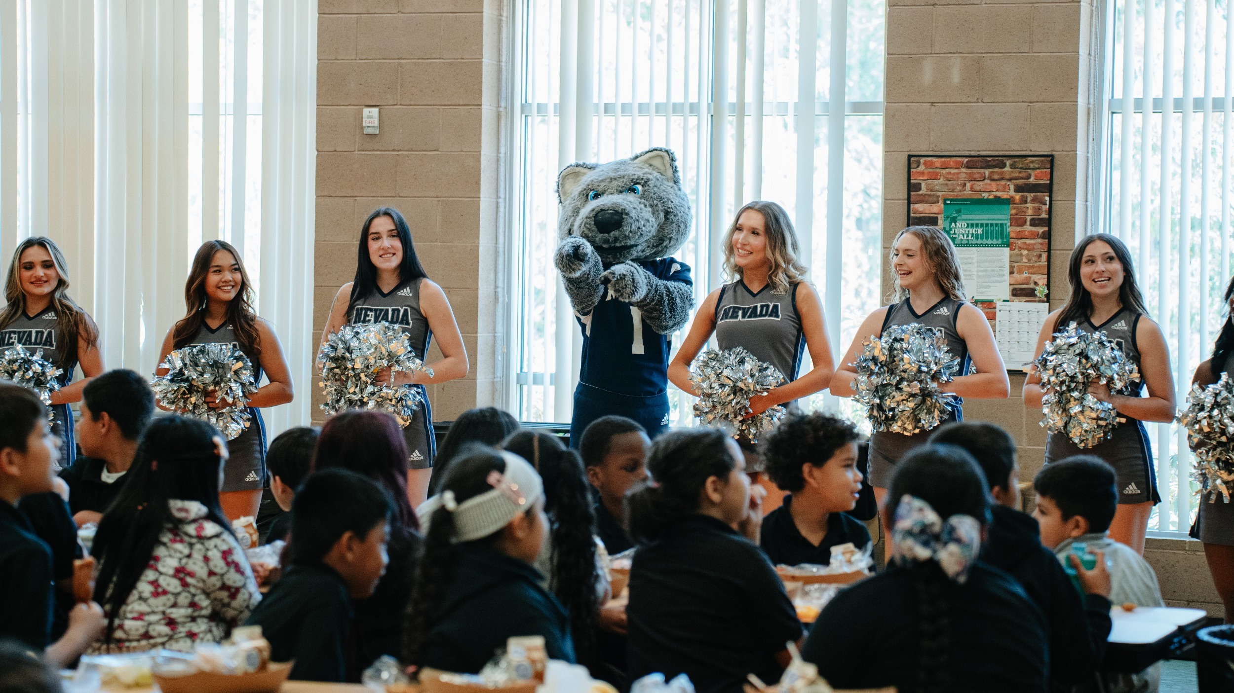 Alphie Jr. mascot pointing at a group of seated schoolchildren with Nevada cheerleaders lined up behind him