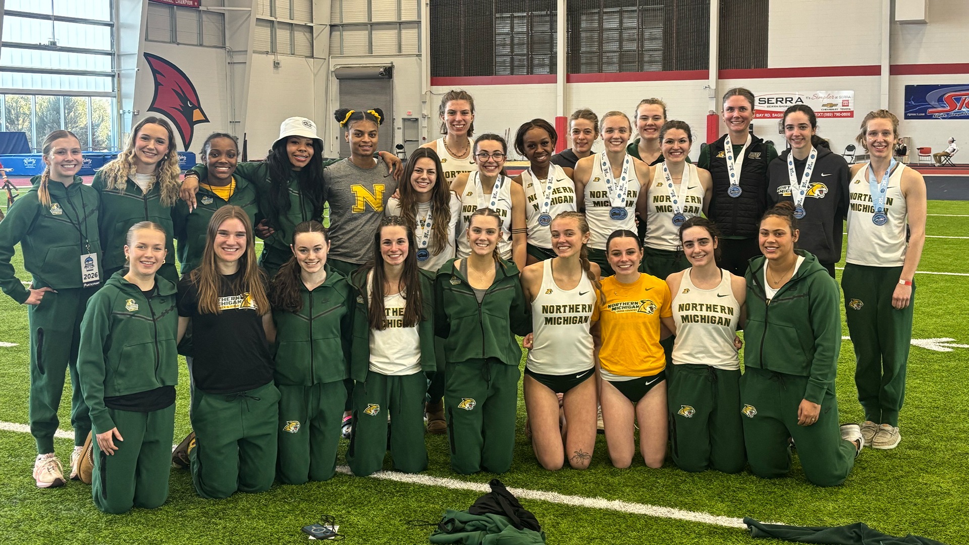 A women's track and field team from Northern Michigan University poses for a group photo on an indoor turf field, some athletes wearing medals.