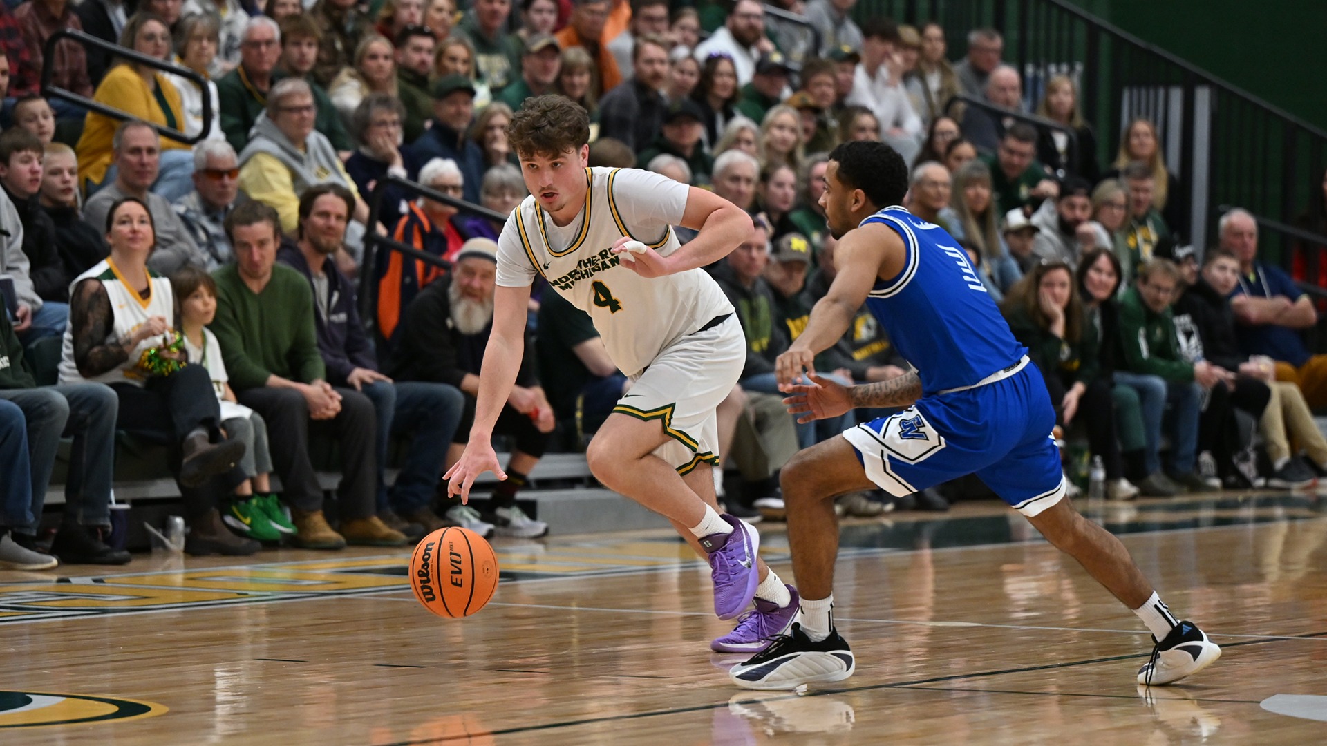 A player in a white #4 jersey dribbles the ball against a defender in a blue jersey. The crowd is full in the background and watches on.