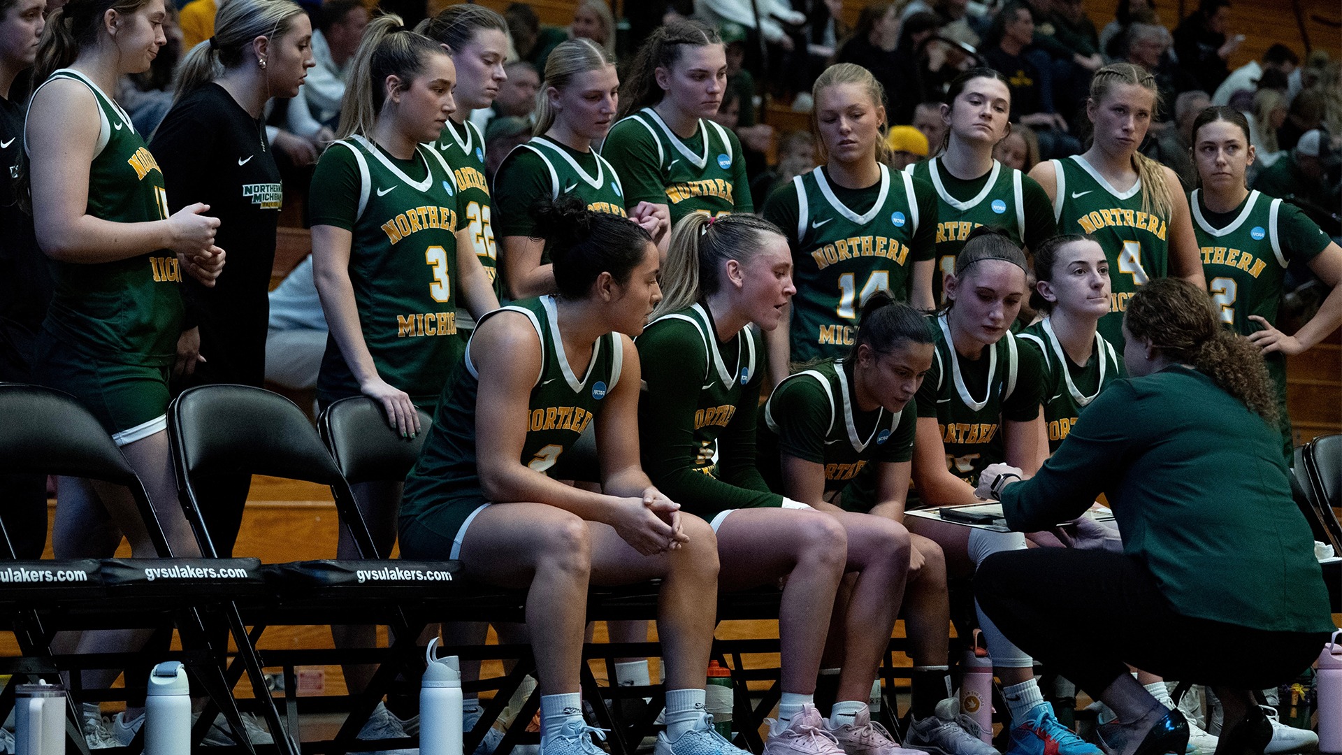 Northern Michigan women’s basketball players gather around their coach during a timeout, sitting and standing along the bench while listening closely to instructions on a clipboard in a crowded gym. Players in green “Northern Michigan” uniforms lean in attentively as fans fill the bleachers behind them.