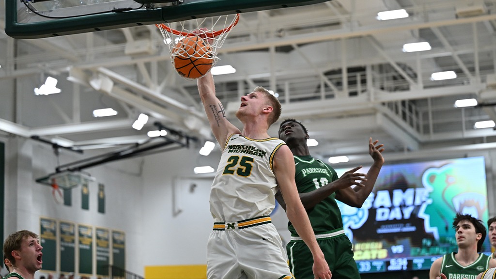 Men’s college basketball action at the rim. A Northern Michigan player in a white No. 25 jersey extends for a one-handed finish as a Parkside defender contests from behind. Players react in the background under bright gym lights.