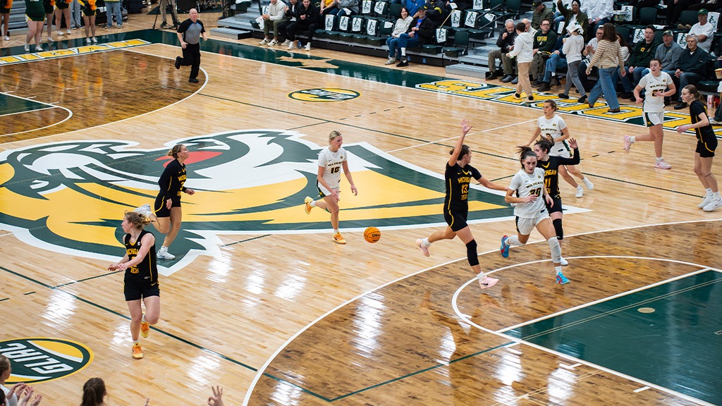 A wide angle of a basketball game, with players and white jerseys and players in black jerseys competing. The crowd looks on and cheers.