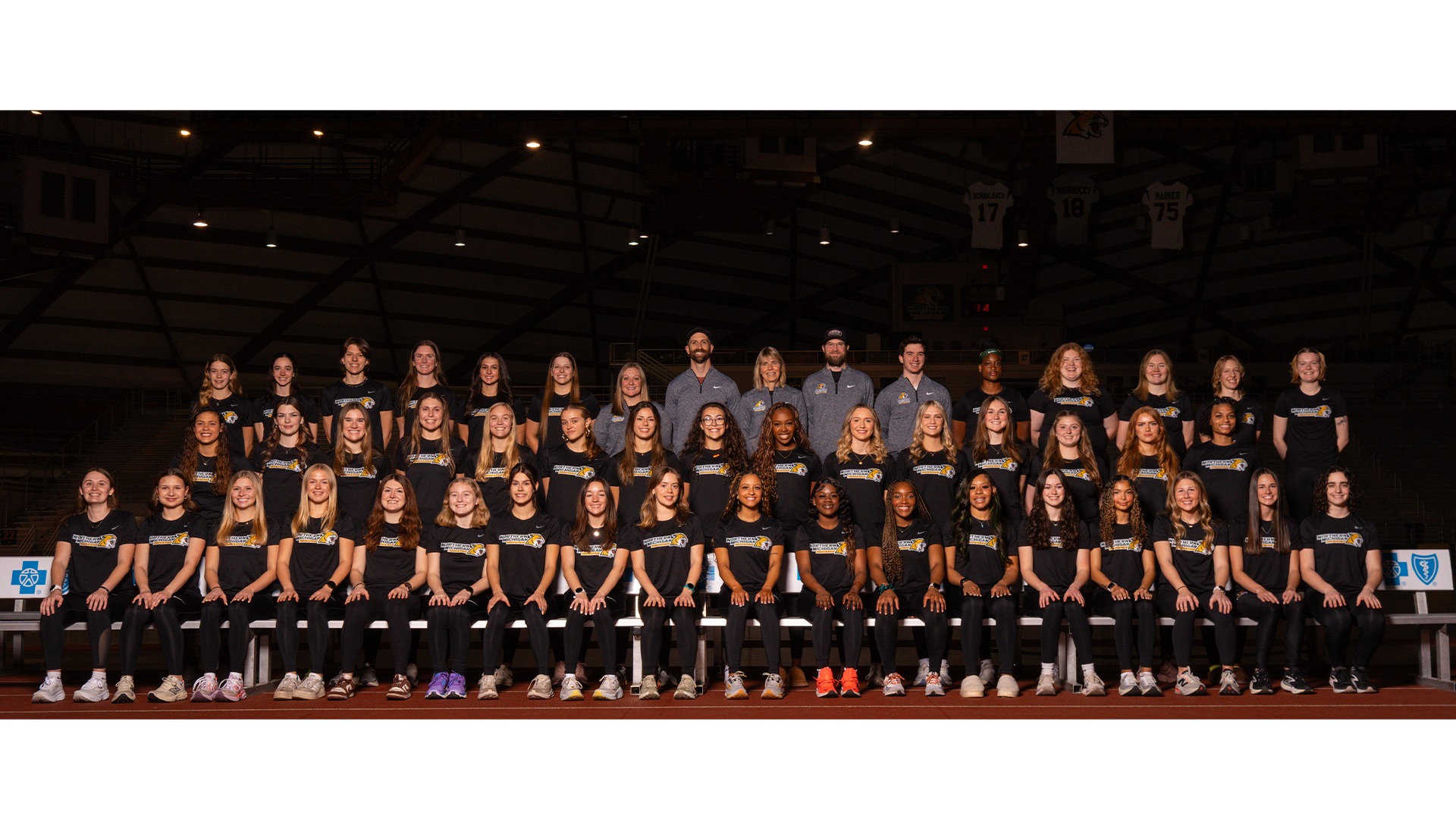 A large group portrait of the Northern Michigan University women's track and field team and coaching staff. The team is posed in three rows inside a large indoor athletic dome. The athletes are wearing matching black short-sleeved shirts with the 