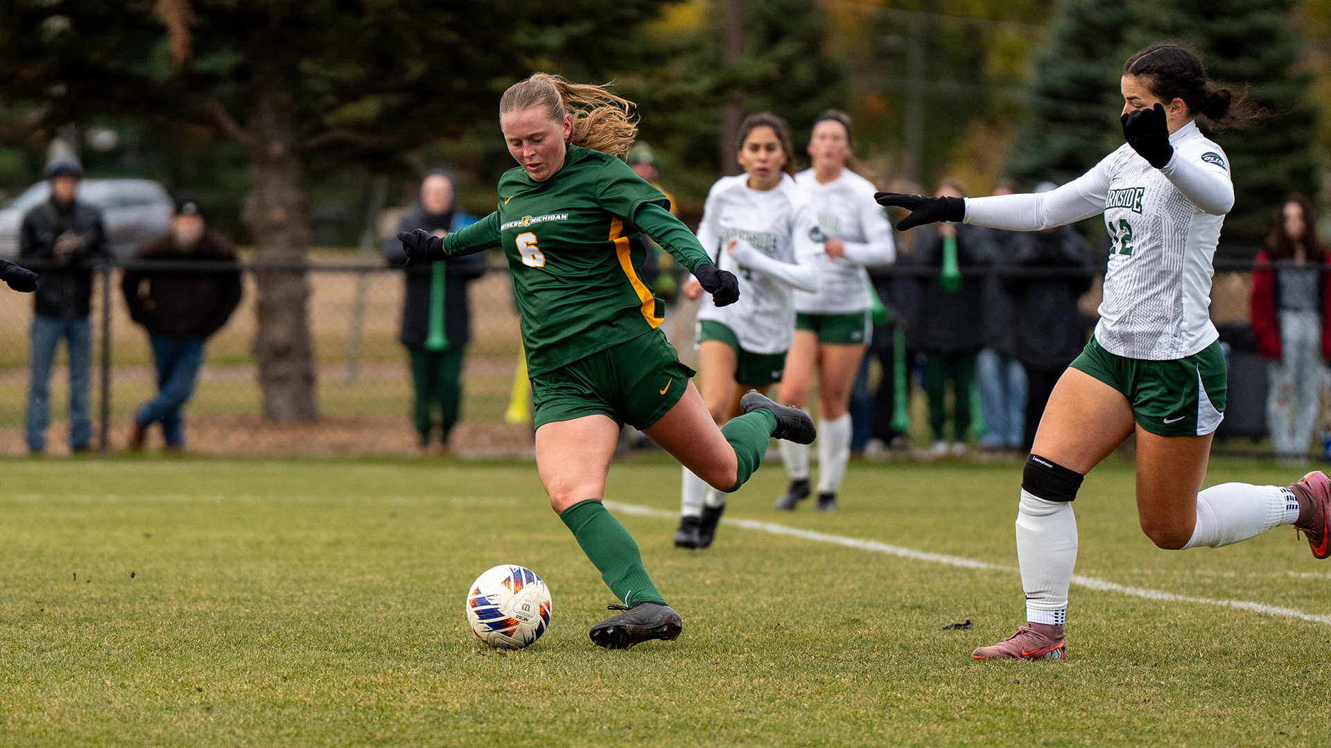 A mid-action shot of the same player (number 6) wearing a long-sleeved green jersey and black gloves. She is captured mid-strike, with her right leg swung back to kick a soccer ball on a grass field. An opposing player in a white Parkside jersey is defending nearby. The background shows spectators and autumn-colored trees behind a fence.