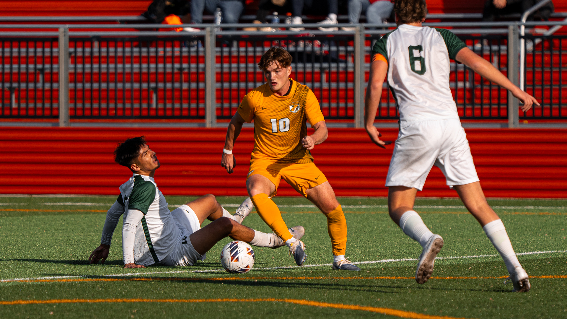 An action shot of the player in his gold number 10 kit, dribbling a soccer ball on a green turf field. One opposing player in a white and green jersey has fallen to the turf in front of him, while another defender (number 6) watches from behind. The background features bright red stadium bleachers.
