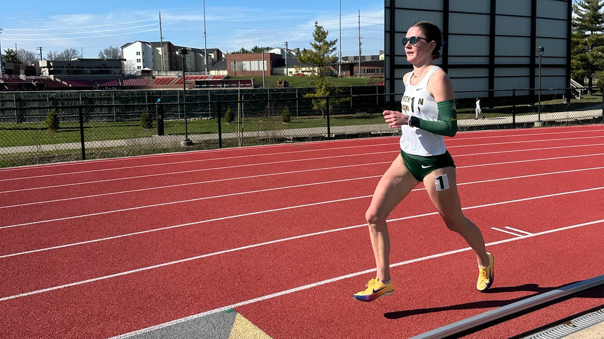 A female runner competing on a red outdoor track under a bright, clear blue sky. She is wearing a white tank top with 