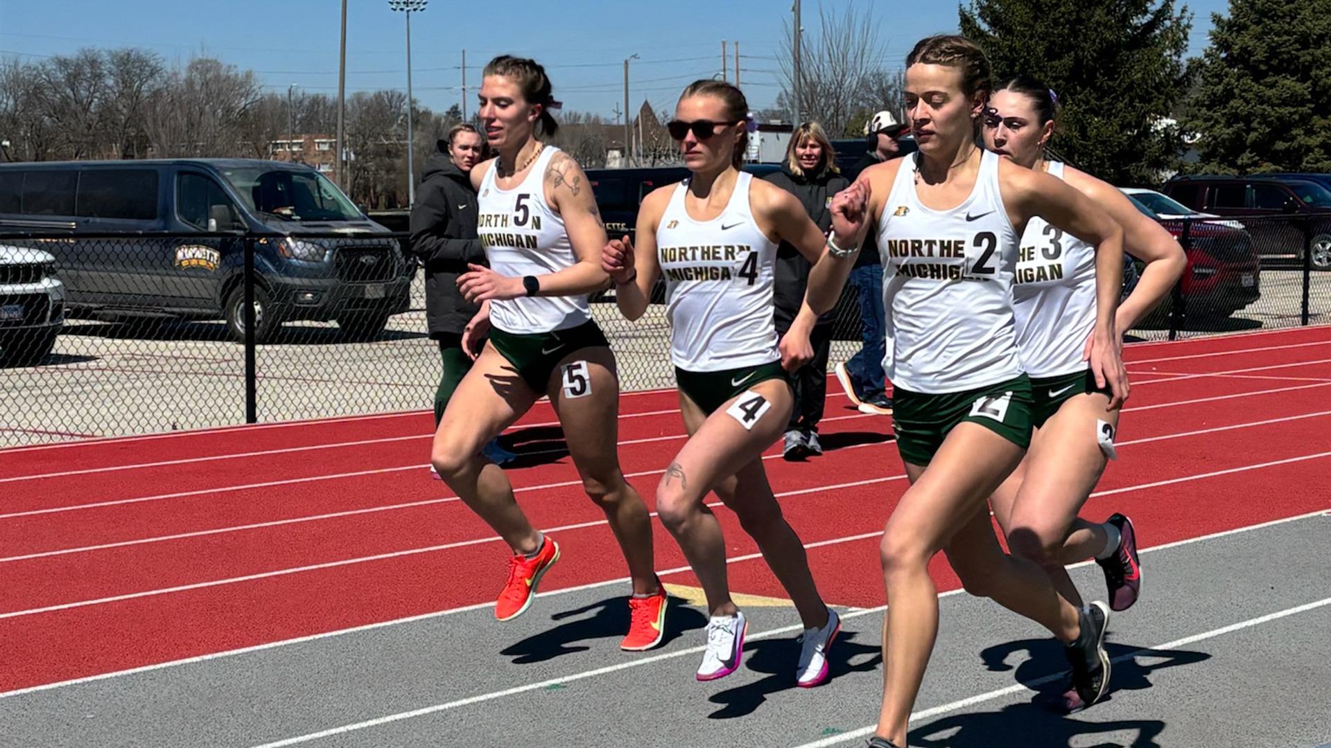  Gemini said Four female track athletes from Northern Michigan are captured mid-stride during a race on a red and grey outdoor track. They are running in a tight pack, all wearing matching white 