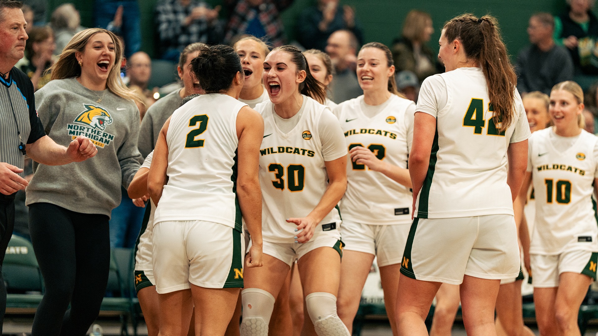 Several players in white jerseys celebrate and embrace afer a play. The fans clap in the background