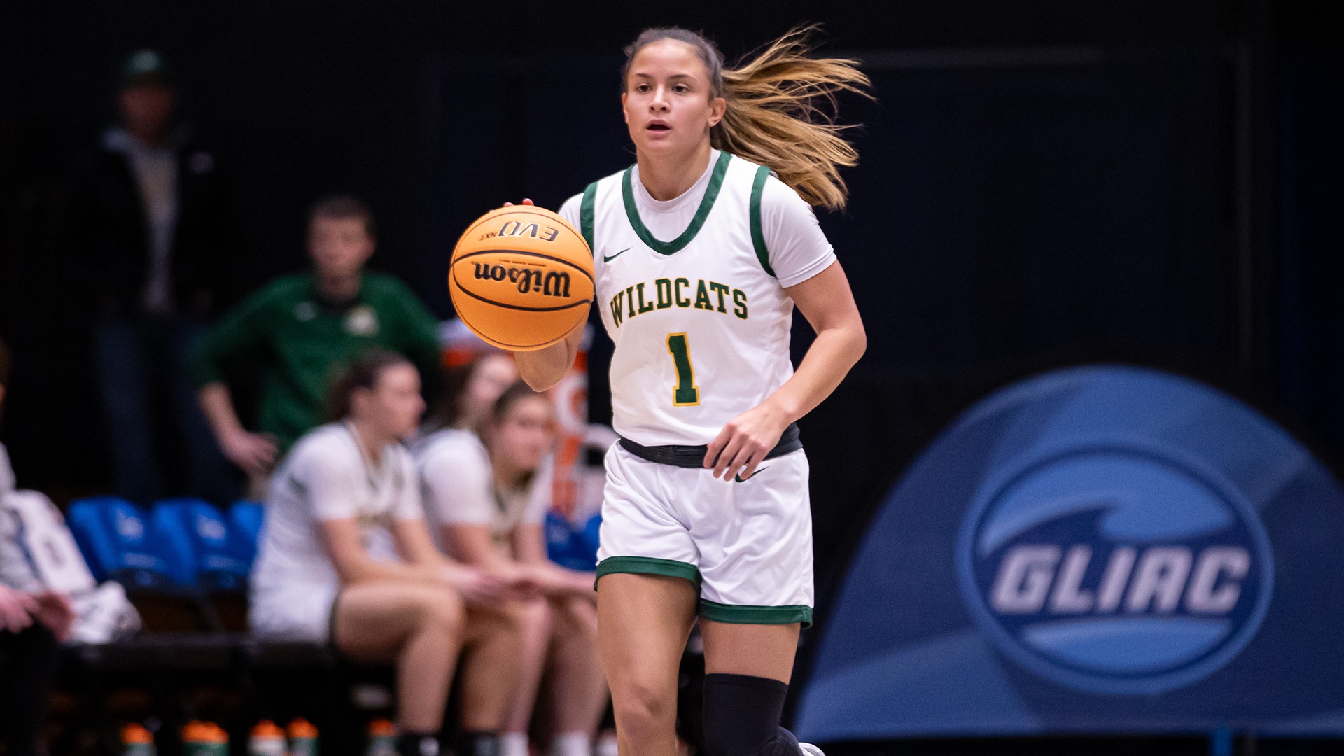 A player in a white #1 jersey dribbles the ball with a dark background and blue GLIAC sign behind her.