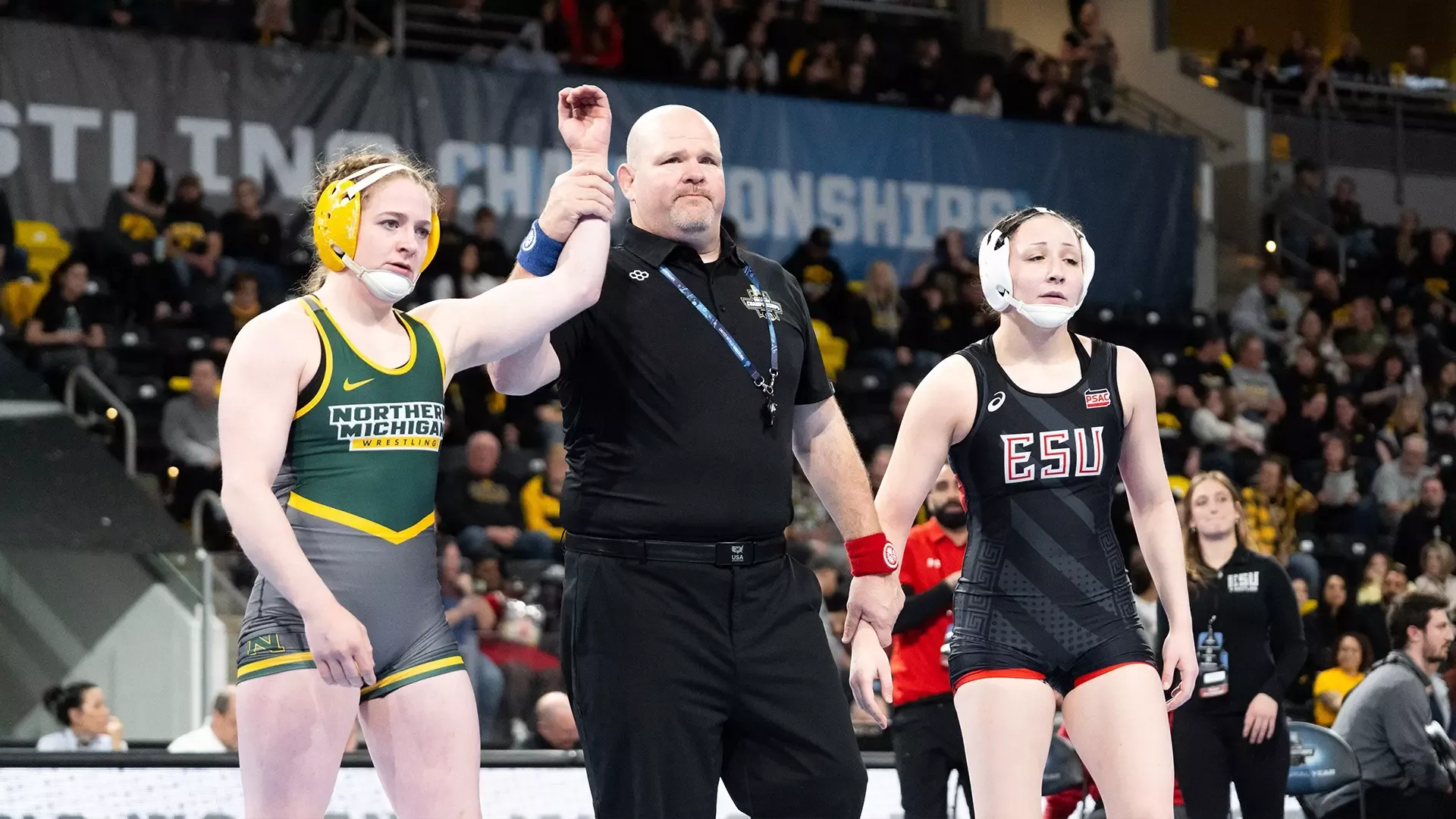 AlisThe image depicts a wrestling match victory ceremony taking place in an arena with an audience. On the left, a wrestler in a green and gray singlet with 