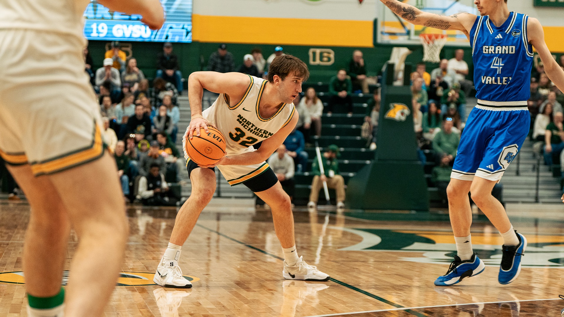 A player in a white #32 jersey bends over with the ball as he looks to make a play against a closing defender in a #4 blue jersey.