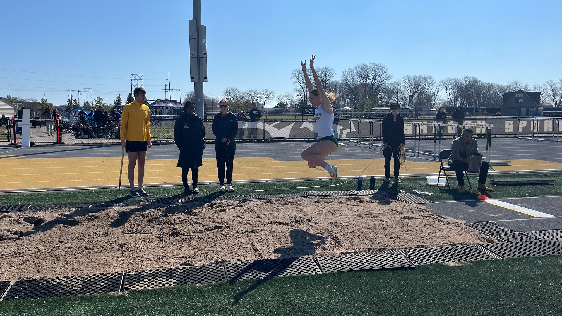 A female track and field athlete is captured in mid-air during a long jump competition on a bright, sunny day. Suspended over a rectangular sand pit, she has her arms extended high and her knees tucked in a classic flight phase, casting a distinct shadow on the ground below. The scene is set on a black and yellow synthetic track, where officials and coaches—including a man in a yellow jacket holding a rake and an official with a clipboard—watch her progress intently. The background features a clear blue sky, bare trees, and track equipment like hurdles, creating a professional and energetic atmosphere for the event.
