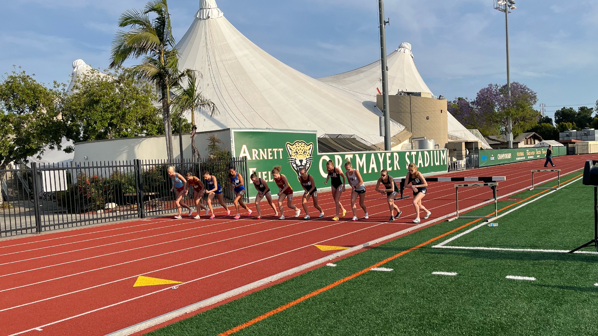 A medium-long outdoor photograph captures ten female student-athletes lined up on an all-weather red running track, crouched at the starting line in their athletic uniforms and bibs. Behind them, a distinctive green field wall features a jaguar logo and reads 
