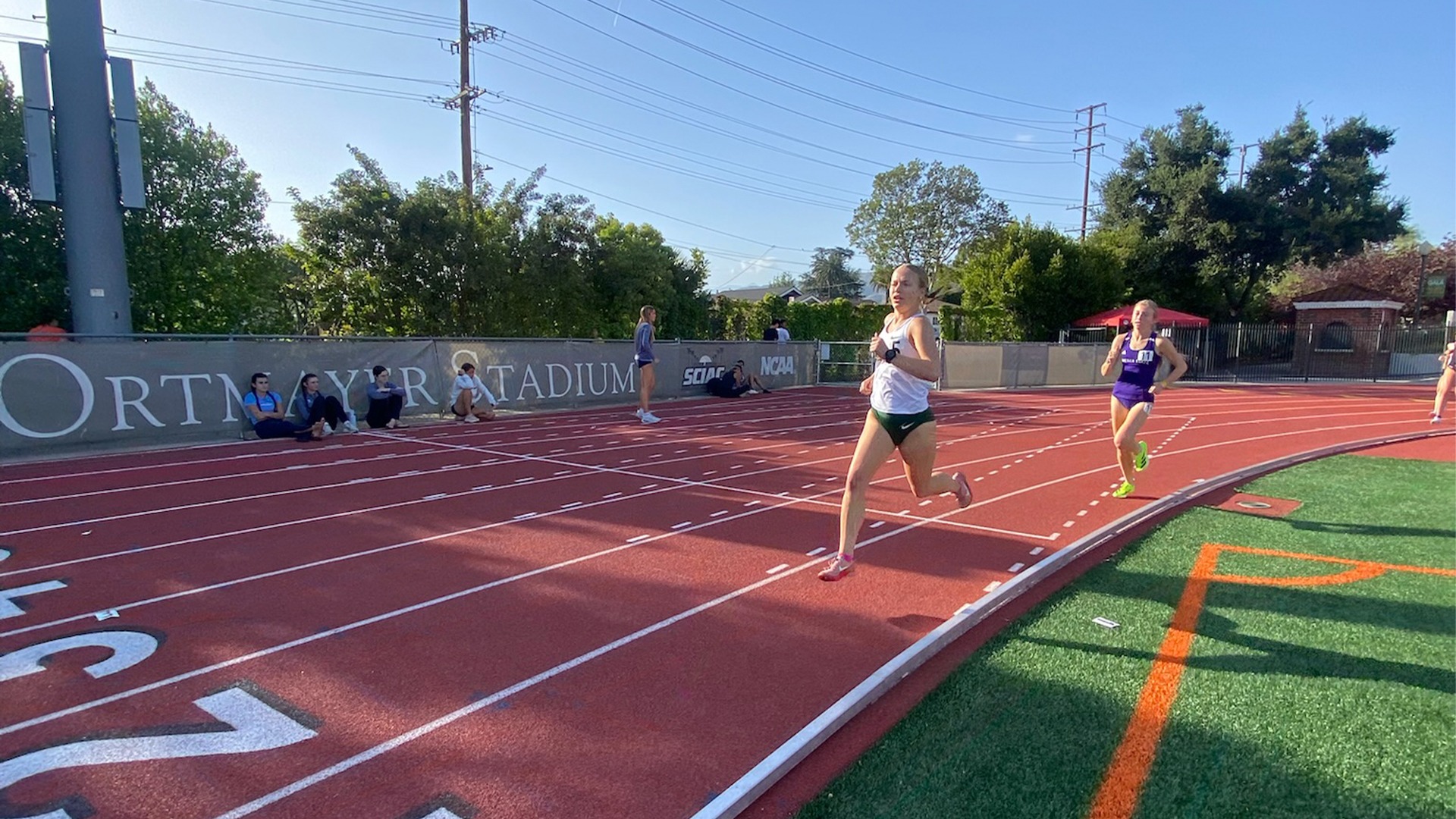  A wide-angle, eye-level shot captures a collegiate track and field event at Ortmayer Stadium on a bright, sunny day. In the foreground, a female runner in a white tank top and dark green shorts is captured mid-stride in the second lane of a red all-weather track, her face showing intense focus as she nears a turn. Closely following her in the third lane is another runner wearing a purple uniform. The long shadows cast across the lanes suggest the late afternoon sun. In the background, several spectators sit along a gray equipment banner, while lush green trees and power lines sit under a clear blue sky. A patch of green turf with bright orange boundary lines is visible in the bottom right corner of the frame.