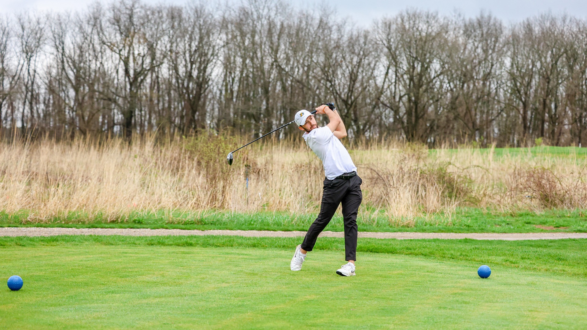A man with a beard and a white baseball cap is shown in the middle of a golf swing on a green tee box. He is wearing a white polo shirt and black pants, and is looking toward the camera as he follows through with his driver. The background features a field of tall, dry grass and a dense line of leafless trees under an overcast sky. Two blue tee markers are visible on the grass in the foreground.