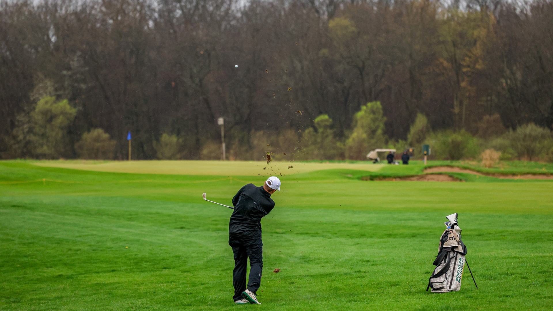 A male golfer in black rain gear and a white hat follows through on a shot from a lush green fairway under a grey, overcast sky. Dirt and grass fly into the air from the impact, and the golf ball is visible mid-flight against the backdrop of a dense, leafless forest. A camouflage golf bag stands on the grass to the right, and a green with a blue flag is visible in the distance.