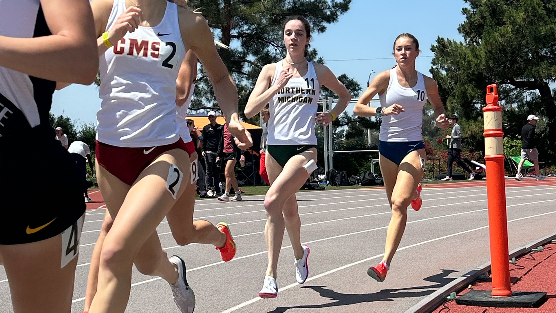  A group of female collegiate athletes competes in a long-distance track race on a clear, sunny day. In the center, a runner wearing a white 