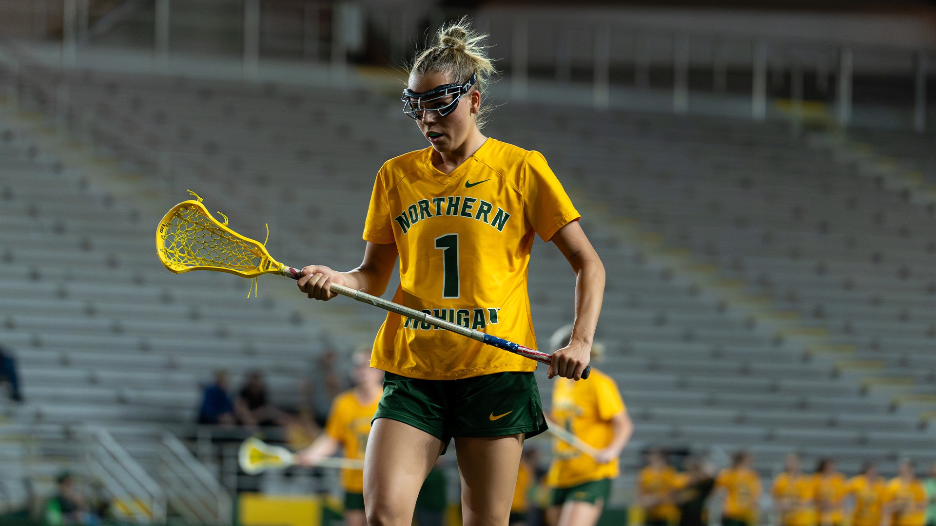 A female lacrosse player for Northern Michigan University stands on a field wearing a yellow jersey with green lettering and the number 1, green shorts, and protective eye goggles. She is holding a lacrosse stick with a yellow head across her body with both hands, looking downward with a focused expression. The background shows blurred stadium seating and other players in similar uniforms.