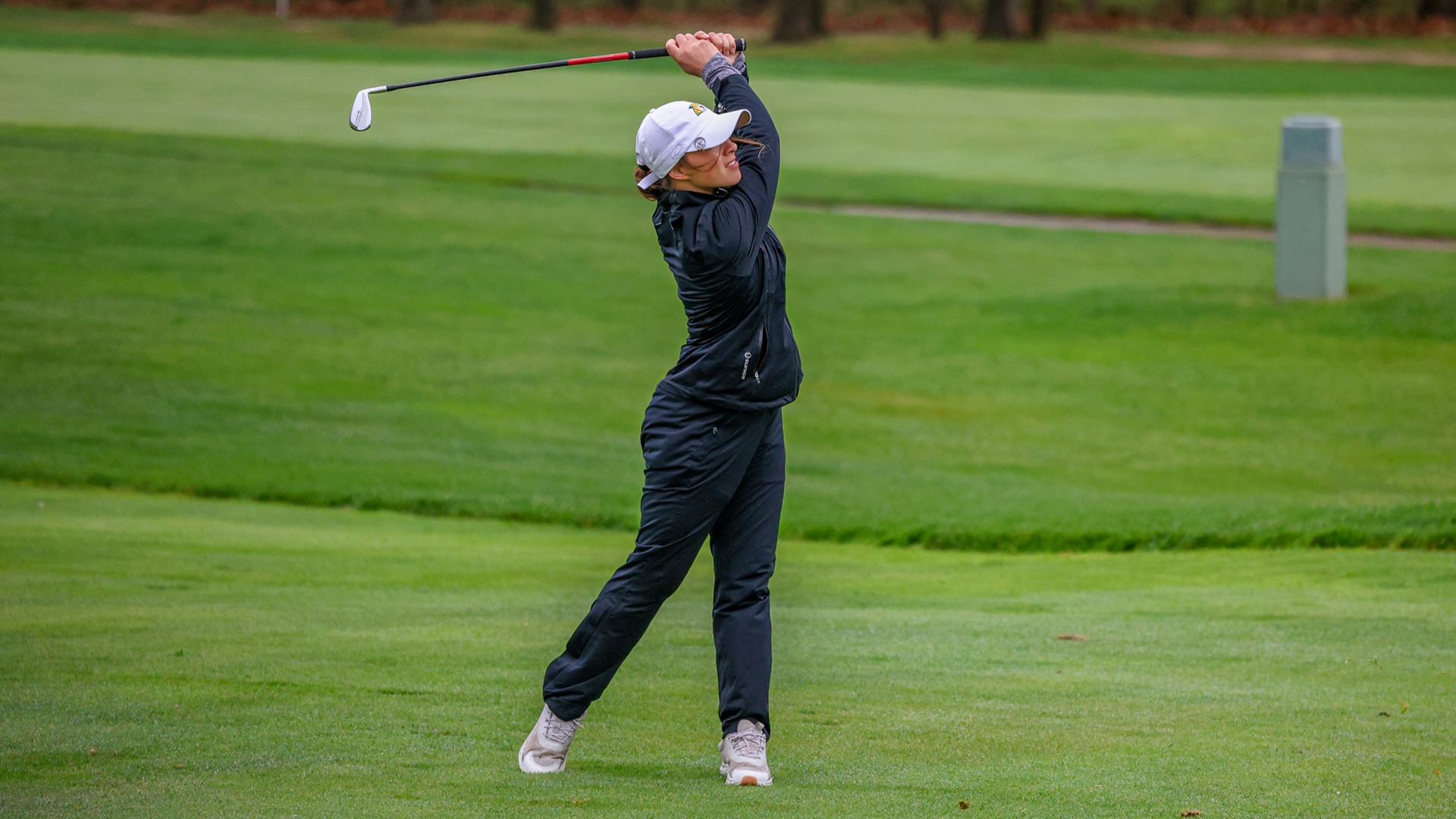  A female golfer in a full follow-through swing on a lush green golf course. She is wearing a white baseball cap, a black long-sleeved athletic jacket, and black trousers. Her body is rotated toward the target, with her weight shifted onto her left foot and her right heel lifted. The golf club is held high over her left shoulder, and she is looking down the fairway. The background features a soft-focus green lawn and distant trees under overcast lighting.