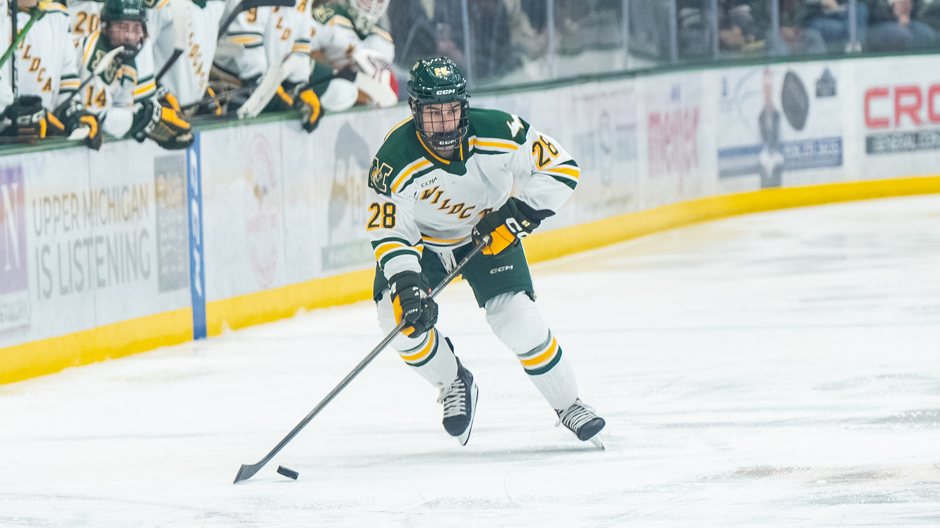 A Northern Michigan University hockey player (number 28) skates across the ice with the puck during a game. He is wearing a white jersey with 
