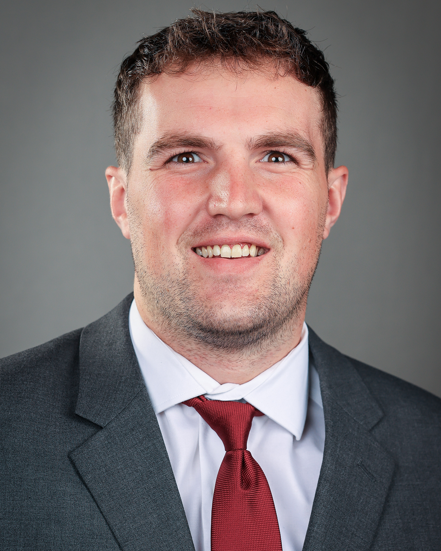 This headshot presents a polished and professional image, characterized by a well-coordinated color palette and approachable body language. The charcoal grey suit provides a sophisticated, neutral base that contrasts effectively with the crisp white shirt, while the deep burgundy tie offers a strong focal point that conveys confidence and energy. Your posture is centered and open, and the genuine, full smile helps project a personality that is both friendly and trustworthy—ideal for making a positive first impression on a professional networking site or a corporate leadership page. The soft, even lighting eliminates harsh shadows, ensuring your features are clear and well-defined against the minimalist grey background.