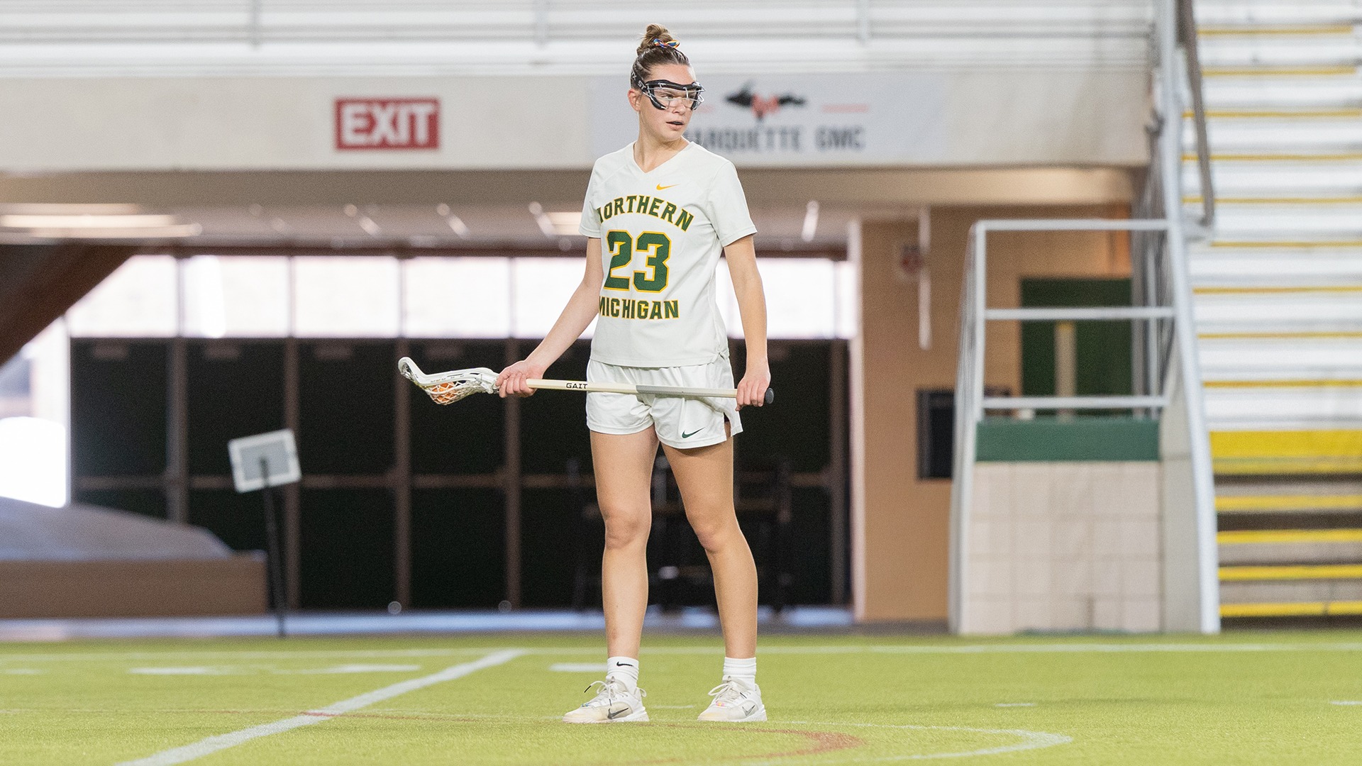 A female lacrosse player for Northern Michigan University stands on an indoor turf field, holding her lacrosse stick horizontally with both hands. She is wearing a white jersey with green and gold lettering that says 