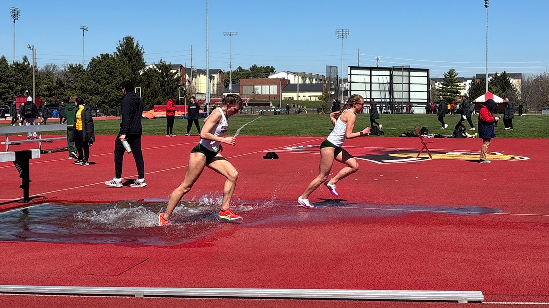 Two female track and field athletes are captured in mid-stride during a steeplechase race on a bright, sunny day. The runner in the foreground, wearing a white tank top and dark green shorts with orange racing spikes, has just landed in the water pit, sending a large splash into the air around her legs. A second runner, dressed in a matching uniform and white spikes, follows closely behind, sprinting across the wet red track surface just past the water jump. The scene is set on a vibrant red all-weather track under a clear blue sky, with other athletes, spectators, and stadium lighting structures visible in the grassy infield and background.