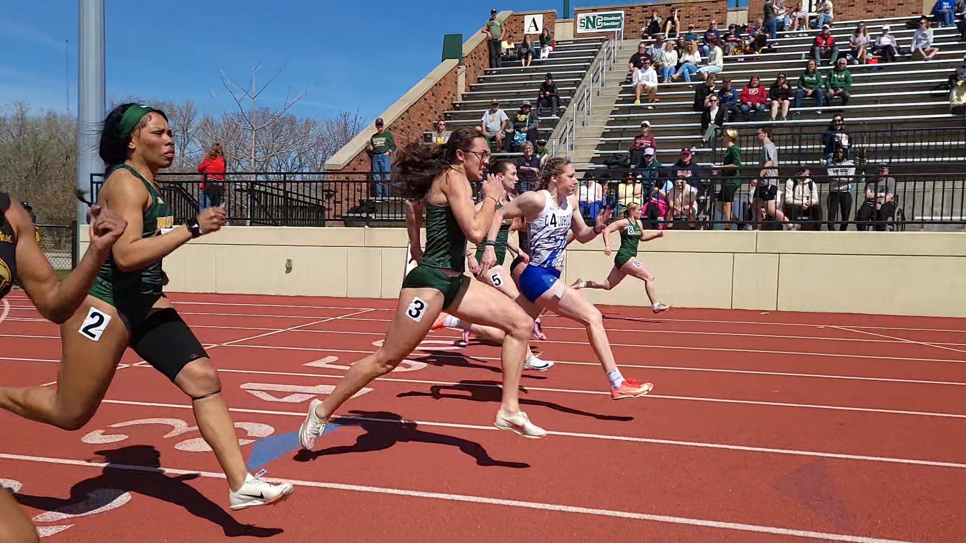 Several female track athletes are captured in a high-intensity sprint during a race on a bright, sunny day. The runners, wearing various collegiate uniforms including green and white and blue and white kits, are spaced across the red all-weather track, their bodies leaned forward in mid-stride with muscles tensed. In the foreground, a runner in a green uniform with the number 2 on her hip leads a pack that includes a competitor in lane 3 and another in a blue-patterned top in lane 4. The background features a concrete wall and a set of bleachers filled with spectators watching from under a clear blue sky.