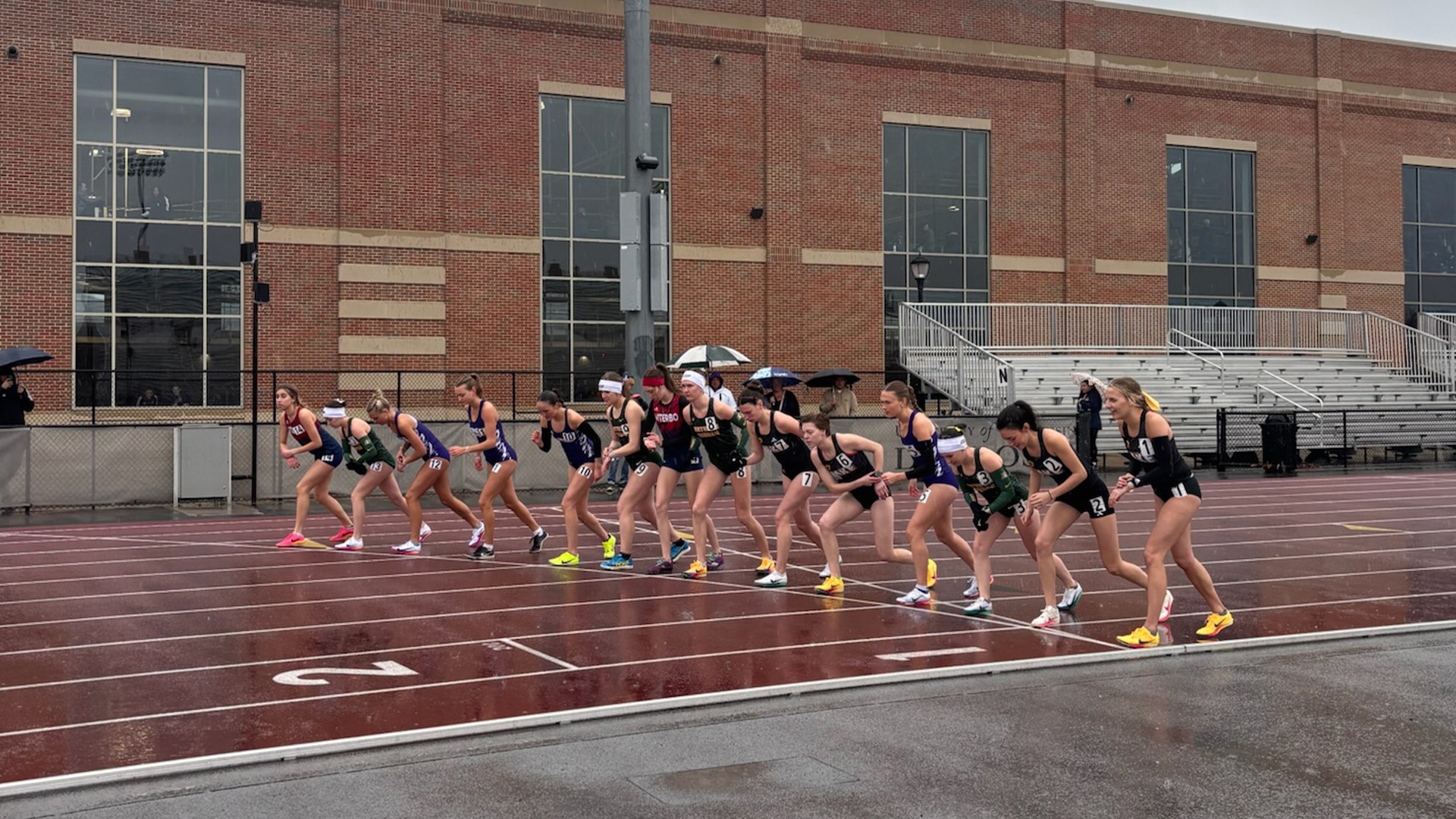 A group of women’s distance runners take the start of a race on a wet outdoor track during rainy conditions, with umbrellas visible in the background and a brick stadium behind them.