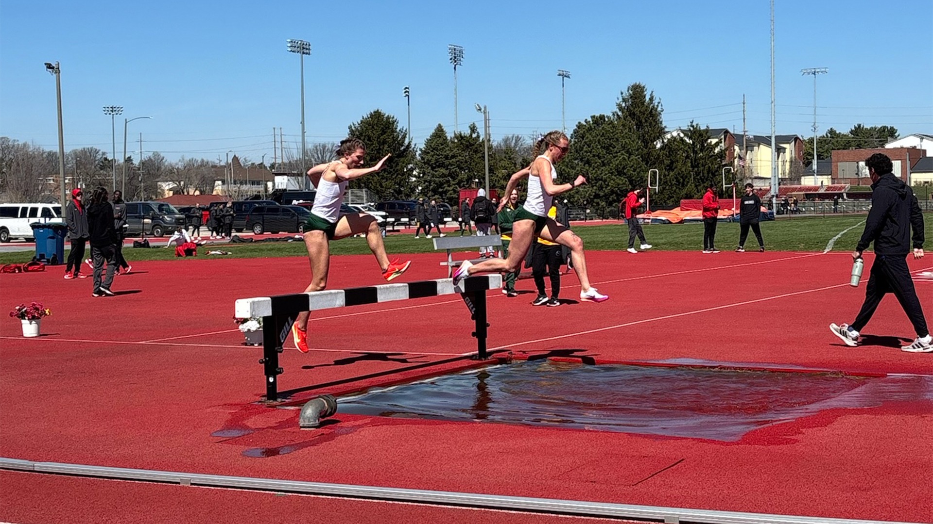  Gemini said Two female athletes are captured mid-air as they jump over a steeplechase hurdle and into a water pit during a track and field event. The runners are wearing white tank tops and green shorts. The athlete in the foreground is leaping off the hurdle with her right foot, while the athlete behind her is just reaching the barrier.  The scene takes place on a bright, sunny day at an outdoor track stadium with a red synthetic surface. In the background, several spectators and other athletes are scattered around the field, and a line of trees and stadium lights are visible under a clear blue sky. A man in a black tracksuit walks along the right side of the track, carrying a water bottle.