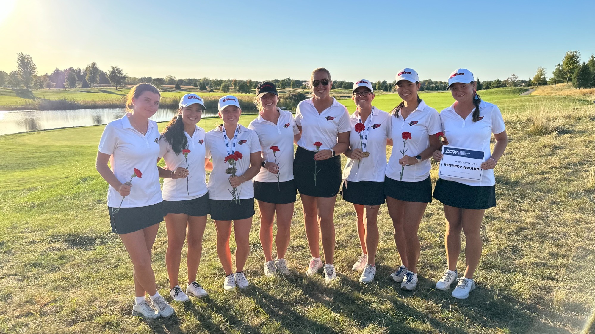 The North Central women's golf poses with their awards in front of a green with water in the background.